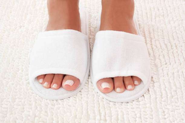 Feet in white slippers on a soft, textured white bath mat, after a shower.