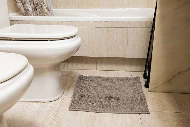 Grey shaggy bath mat on tiled bathroom floor next to two white toilets.