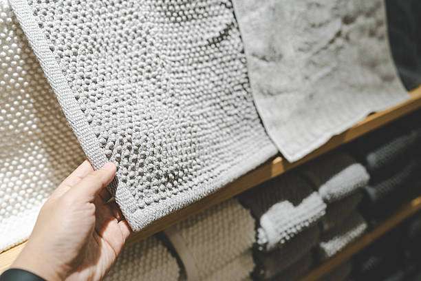 Hand holds a grey textured bath mat, with more mats displayed on shelves.